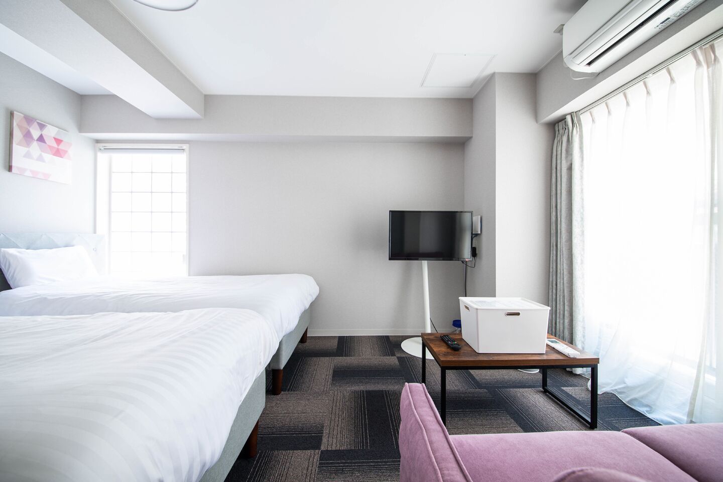 A hotel room with two beds, a flat-screen TV, a small table with a remote control, and a window with curtains.