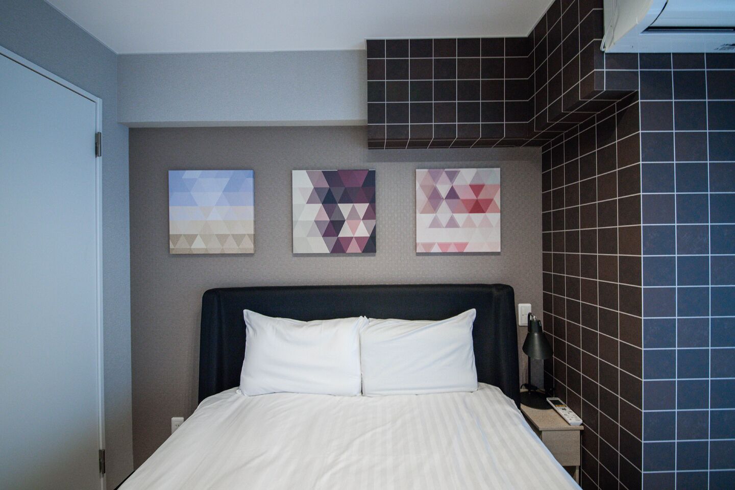 Una habitación de hotel con una cama, una mesita de noche, una lámpara y tres obras de arte en la pared con motivos geométricos.