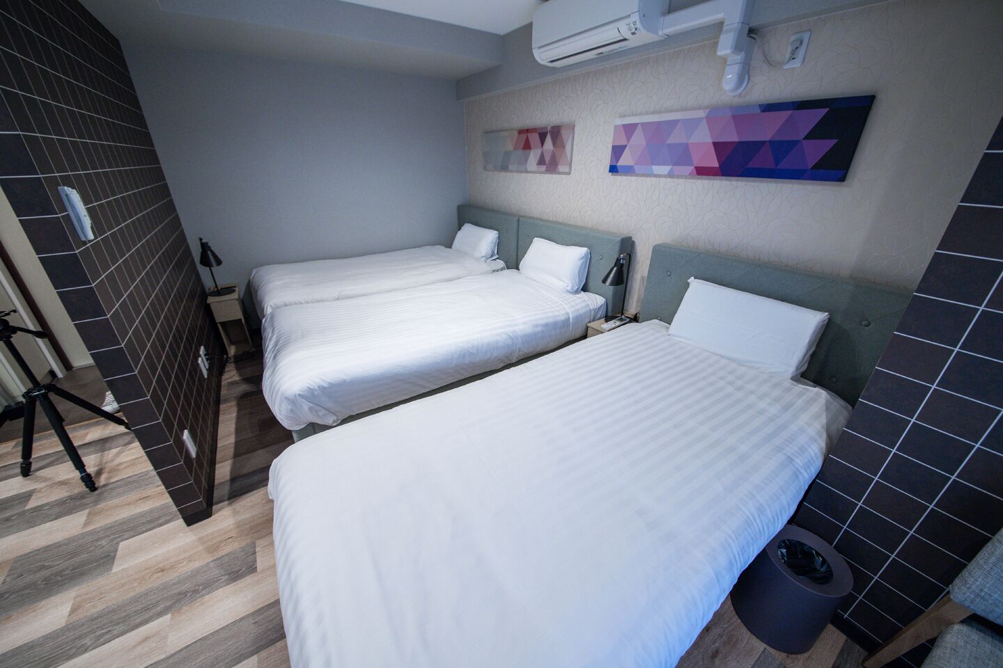 A hotel room with two single beds, a small table, a trash bin, and a wall-mounted air conditioner.