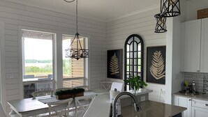 A kitchen with white cabinets, a dining area with white chairs, a large window, and pendant lights. - Your Own Private Waterfront Resort! Pool, Hot Tub, Lagoon, Kayaks, Bikes & More! (Gulf Shores)