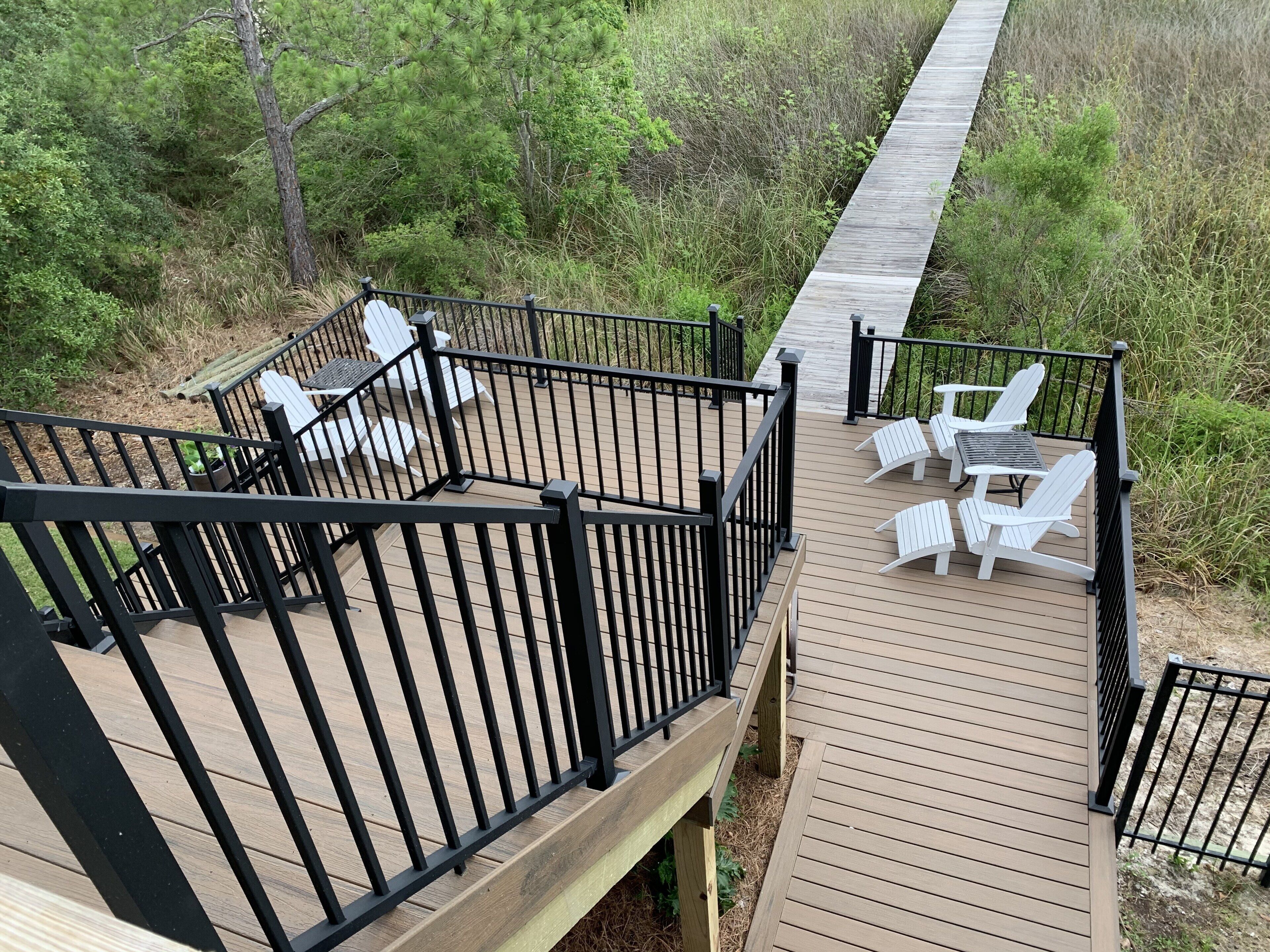 Terraza de madera con sillas y mesas blancas, rodeada por una barandilla negra, con vistas a un paisaje natural.