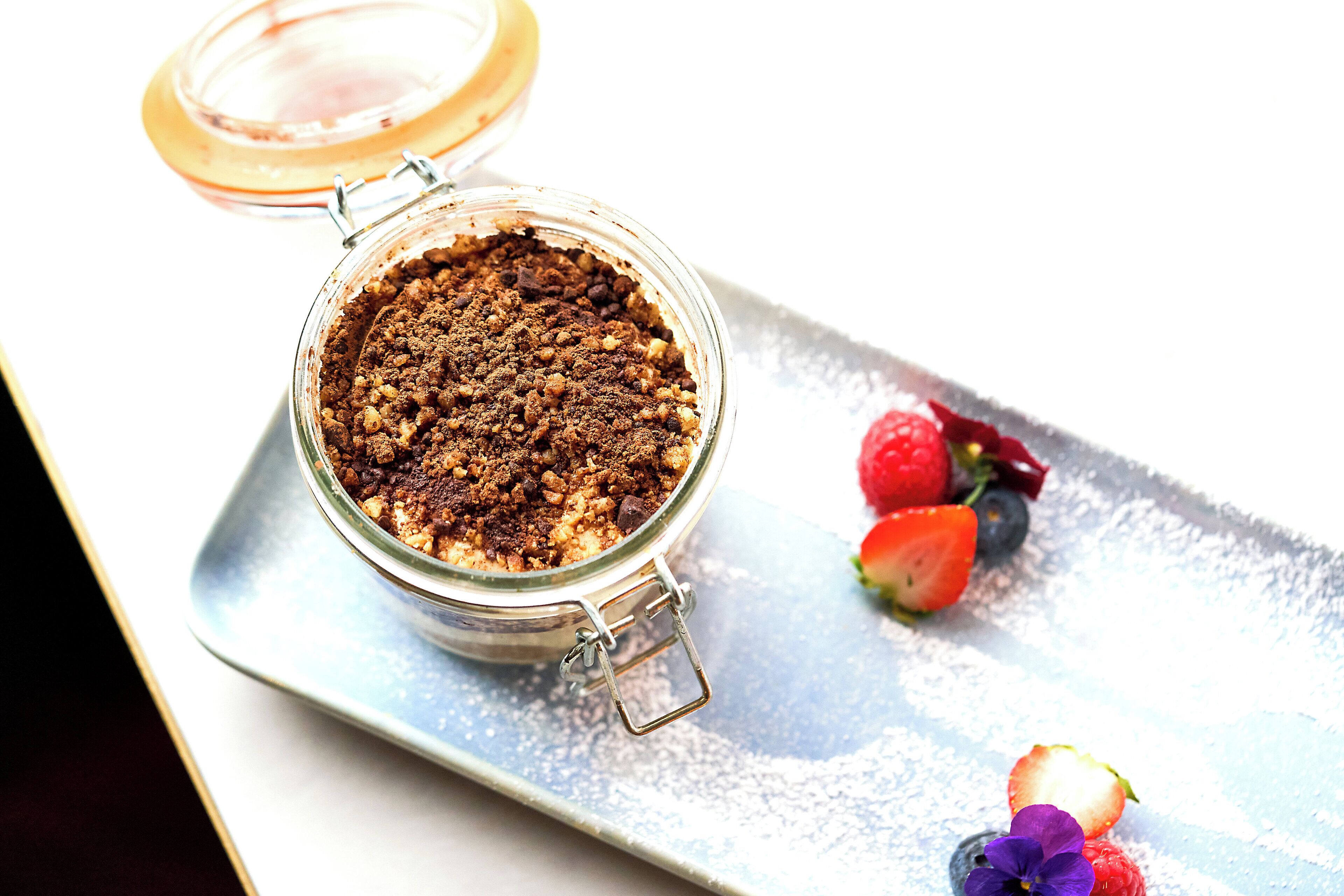 Un dessert servi dans un bocal en verre muni d’un couvercle métallique à fermoir, accompagné de fraises et de framboises présentées sur un plateau en argent.