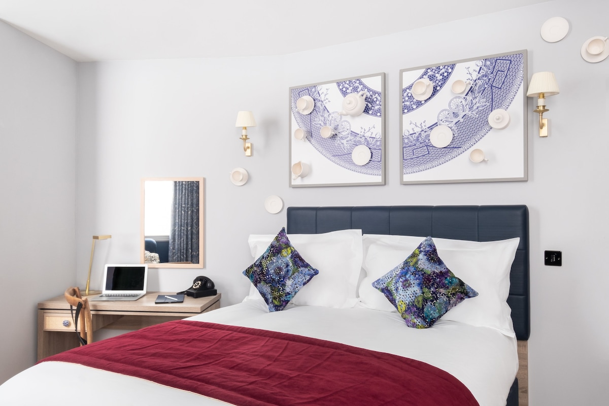 A hotel room with a bed, a desk with a laptop, a phone, and a lamp. There are two wall-mounted lamps and a framed artwork above the bed.