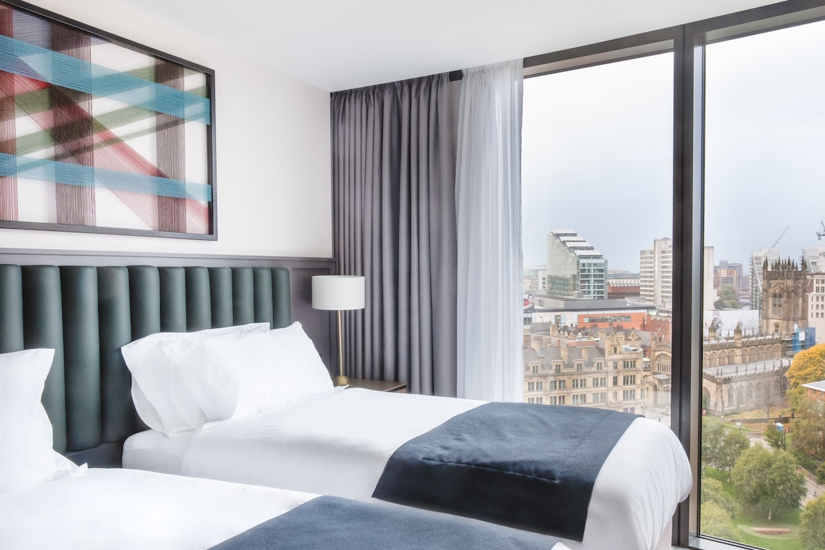 A hotel room with a large window, a bed with white and dark blue bedding, a modern headboard, and a city view.