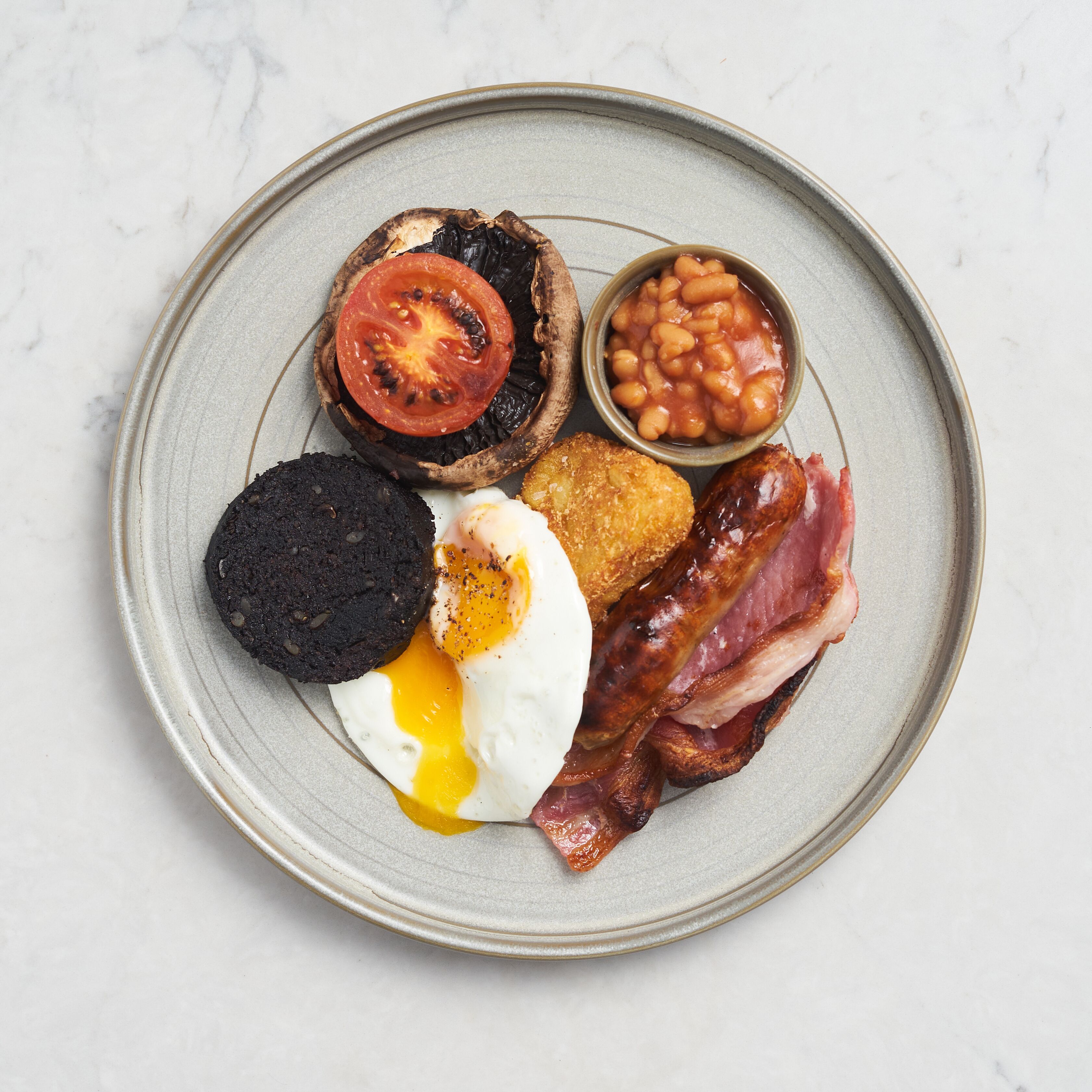A plate of breakfast food including eggs, bacon, beans, a tomato slice, and a black pudding.