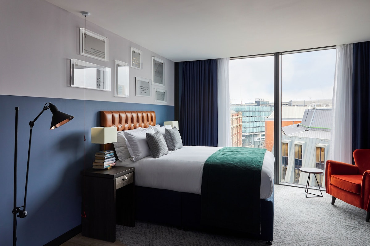 A hotel room with a bed, a desk, a chair, a lamp, and a view of the city through large windows.