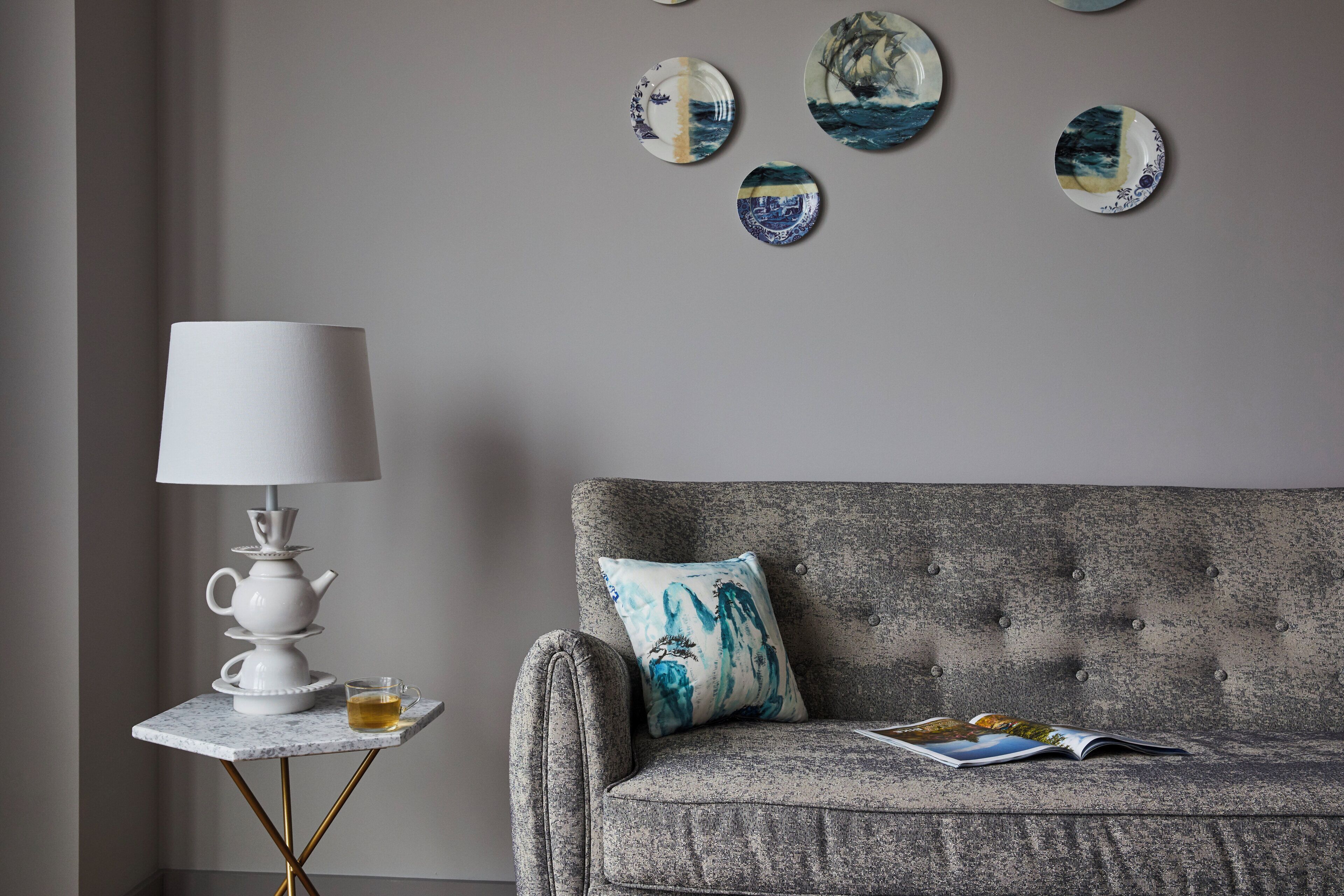 A room with a grey sofa, a white table lamp, a small table with a teapot and cup, and a magazine on the sofa.