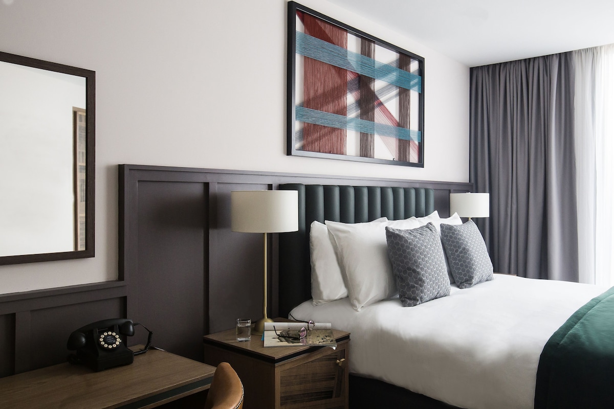 Une chambre d’hôtel comprenant un lit, deux lampes de chevet, un bureau avec un téléphone et une œuvre d’art encadrée au-dessus du lit.