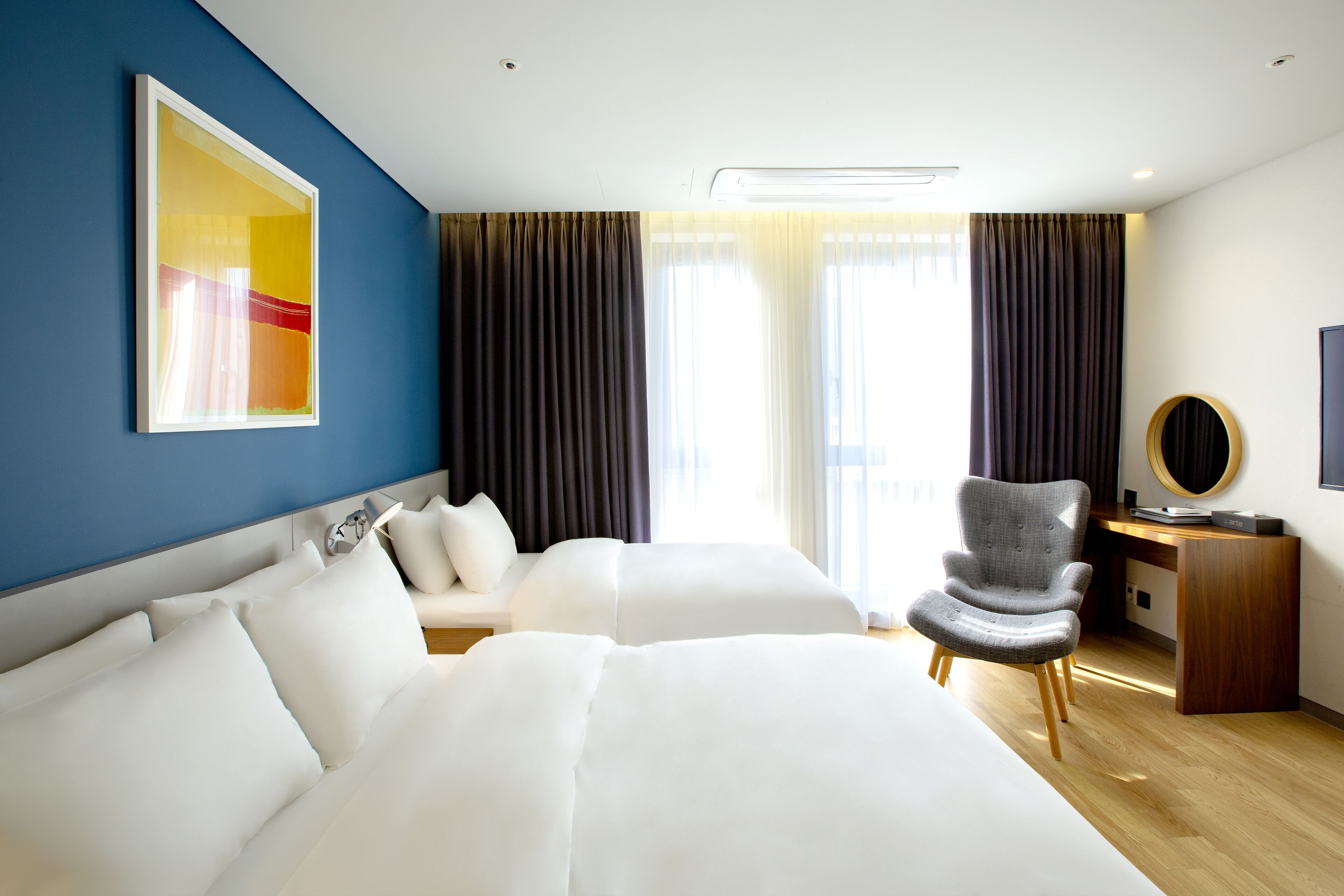 Une chambre d’hôtel avec deux lits, un bureau, une chaise, une grande fenêtre avec des rideaux et une œuvre d’art accrochée au mur.