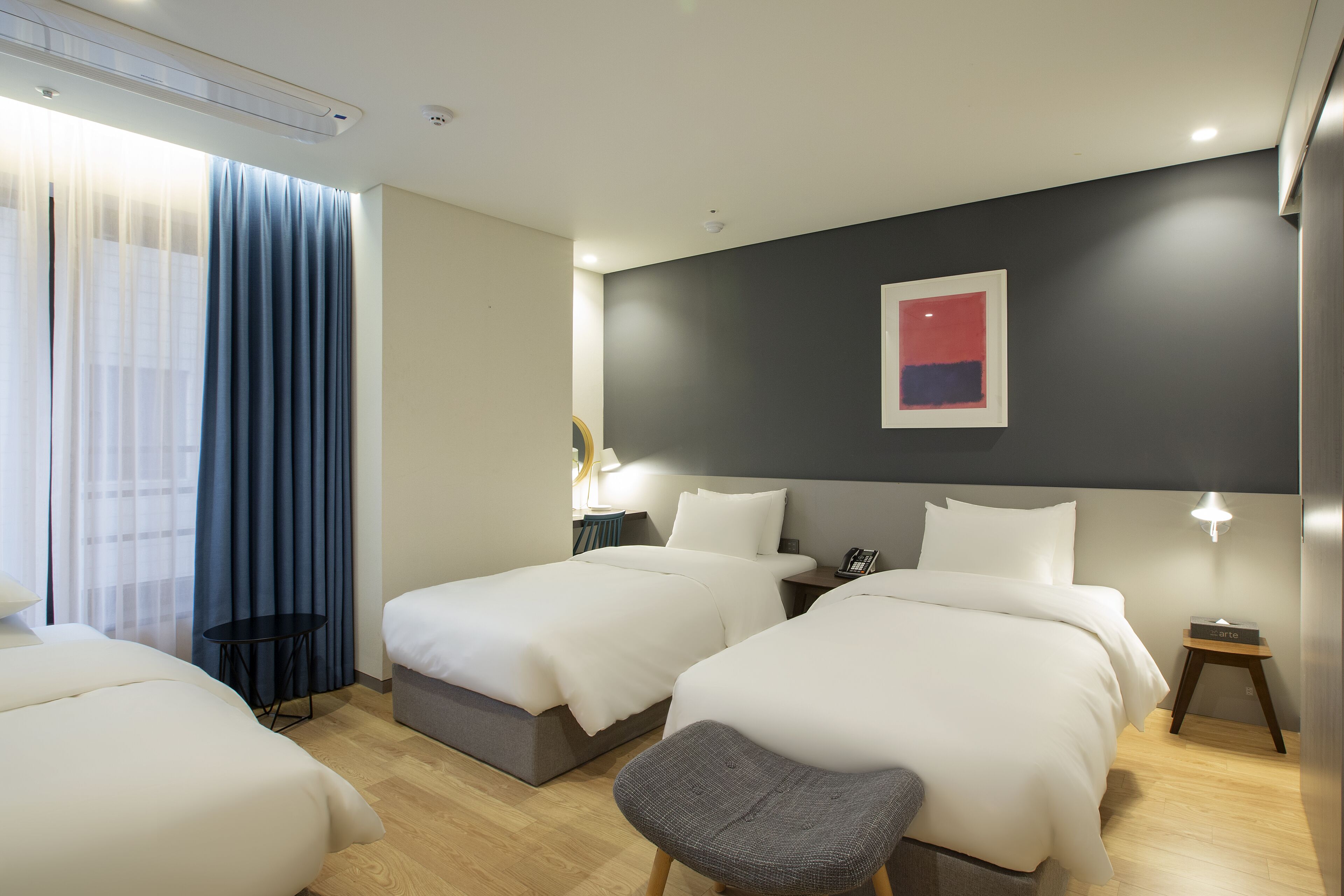 A hotel room with two beds, a small table, a chair, a bedside table with a lamp, and a painting on the wall.