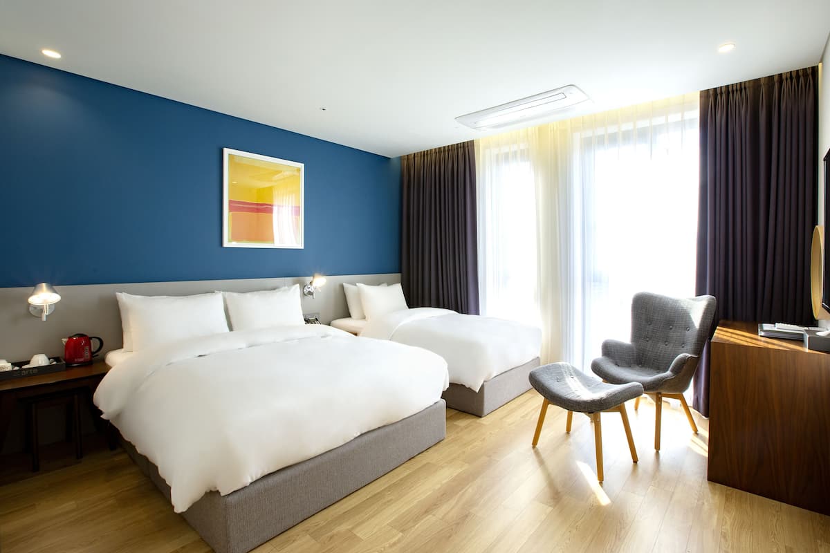 Habitación de hotel con dos camas, un escritorio, una silla y un ventanal grande con cortinas.