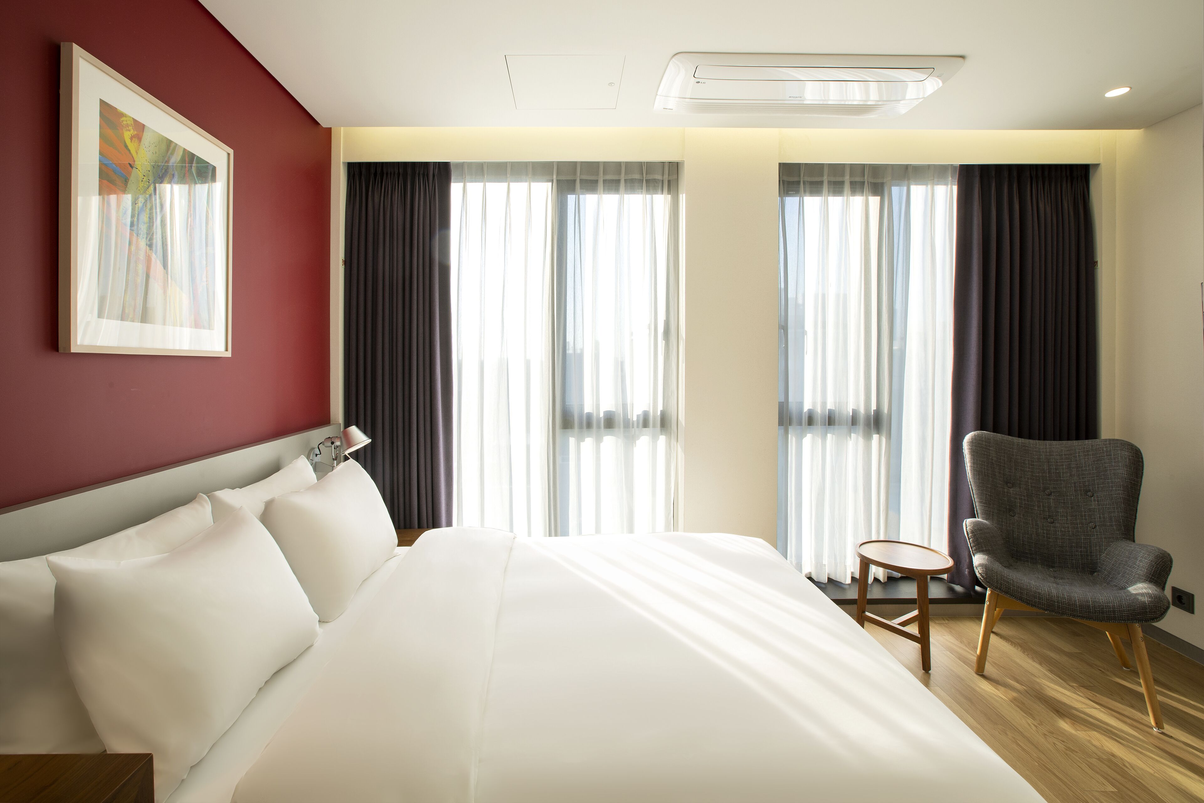 Une chambre d’hôtel avec un grand lit, une chaise et une petite table. Il y a des rideaux aux fenêtres et un tableau au mur.