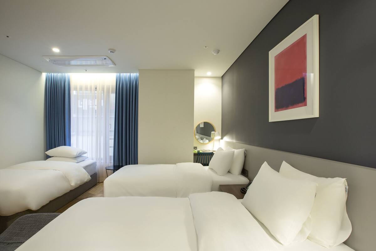 Habitación de hotel con dos camas, un ventanal con cortinas azules, un espejo redondo y un cuadro abstracto rojo en la pared.