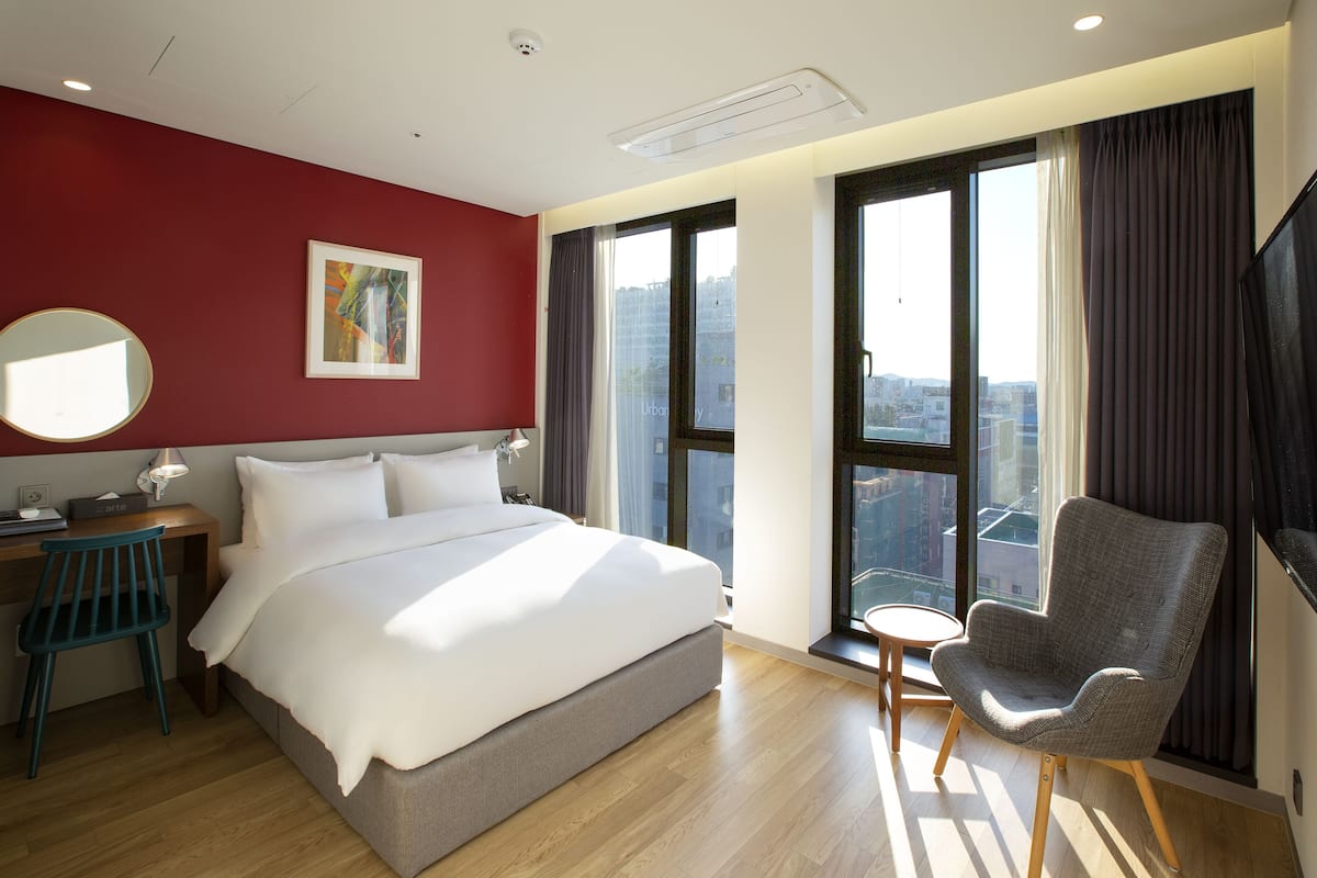 Una habitación de hotel moderna con una cama grande, un escritorio, una silla y un ventanal con vistas a la ciudad.