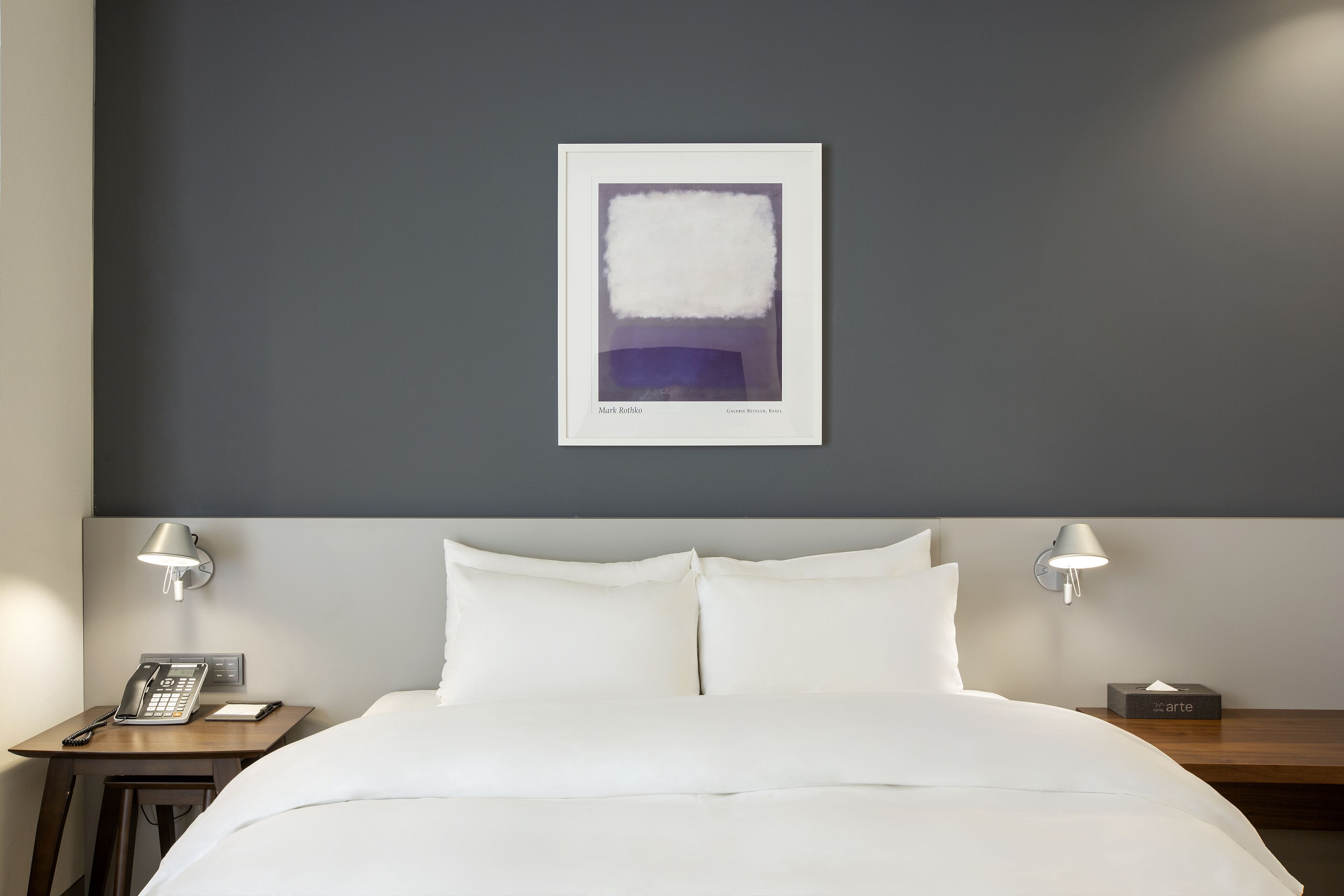 A hotel room with a bed, two bedside tables, a wall-mounted artwork, and a bedside lamp.