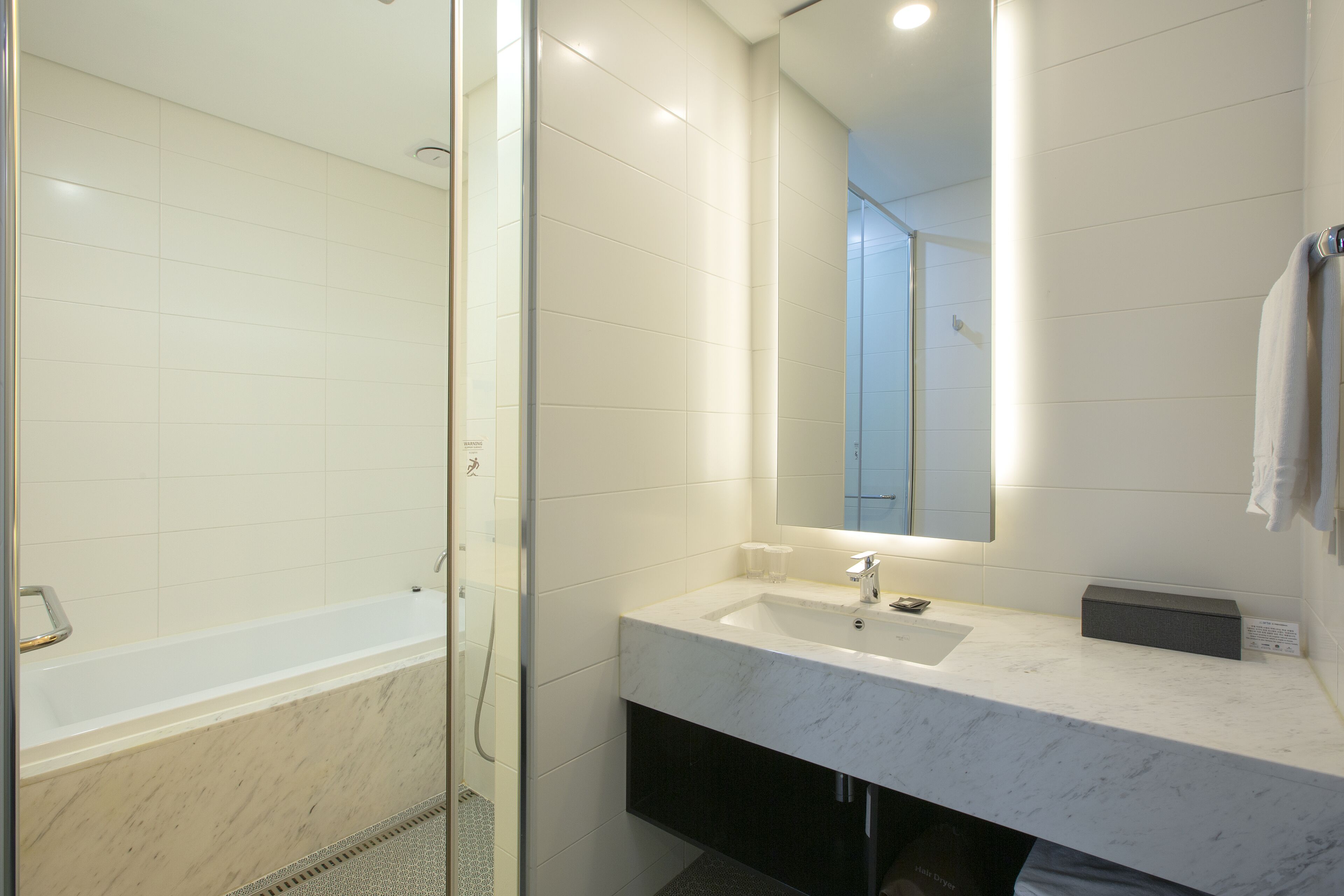 Un baño moderno con ducha de vidrio, lavamanos blanco sobre encimera de mármol y un espejo grande.