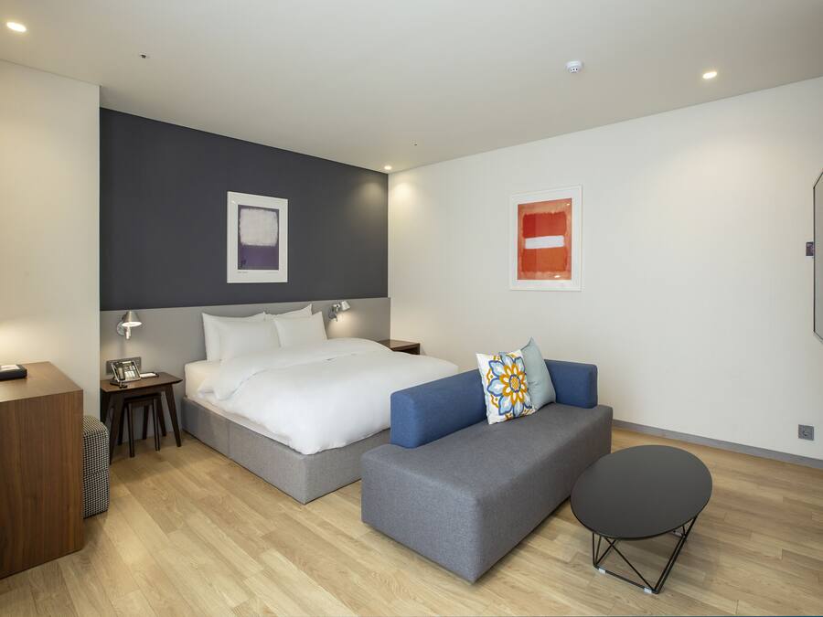 A modern hotel room with a large bed, a sofa, a small table, and a flat-screen TV mounted on the wall.