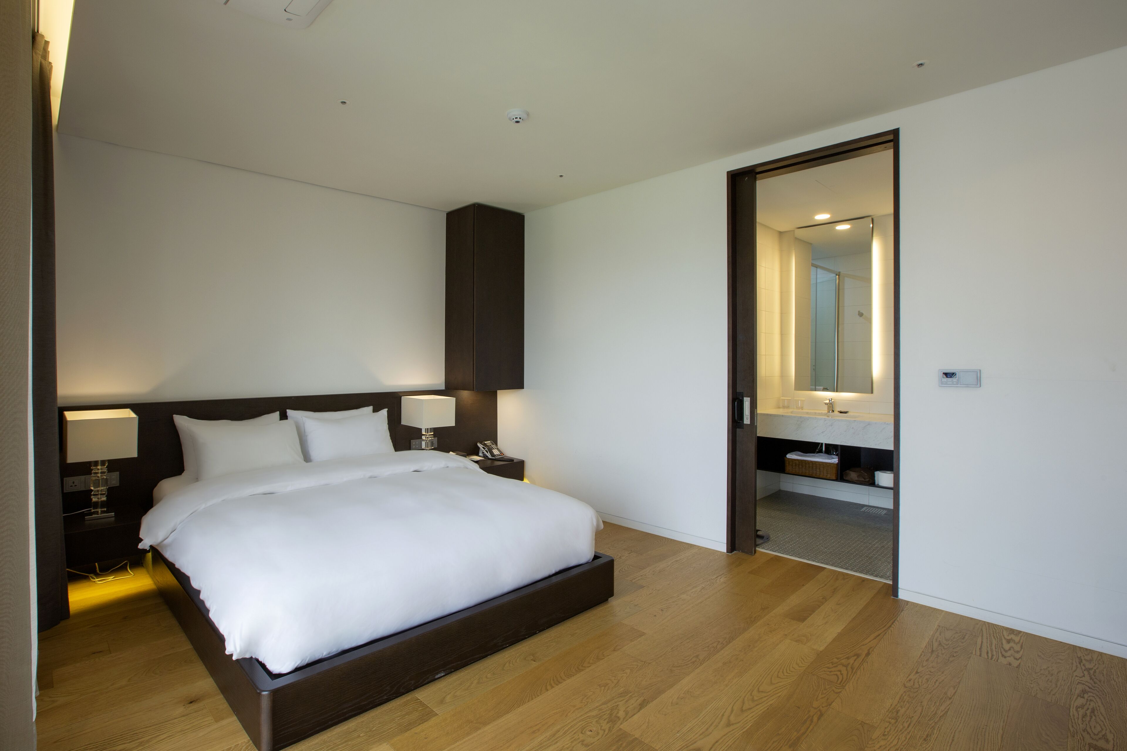 A modern hotel bedroom with a large bed, bedside lamps, a dark headboard, and a wooden floor.