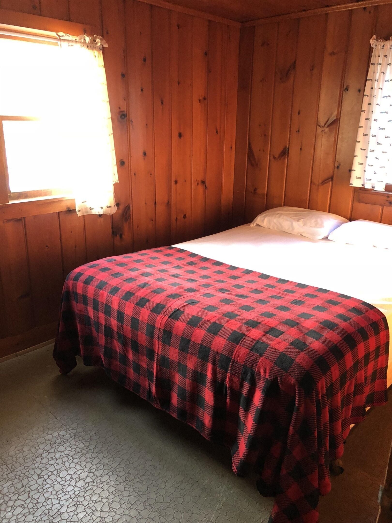 Una habitación con paredes de madera, una cama con colcha de cuadros rojos y negros, y una ventana con cortina blanca.
