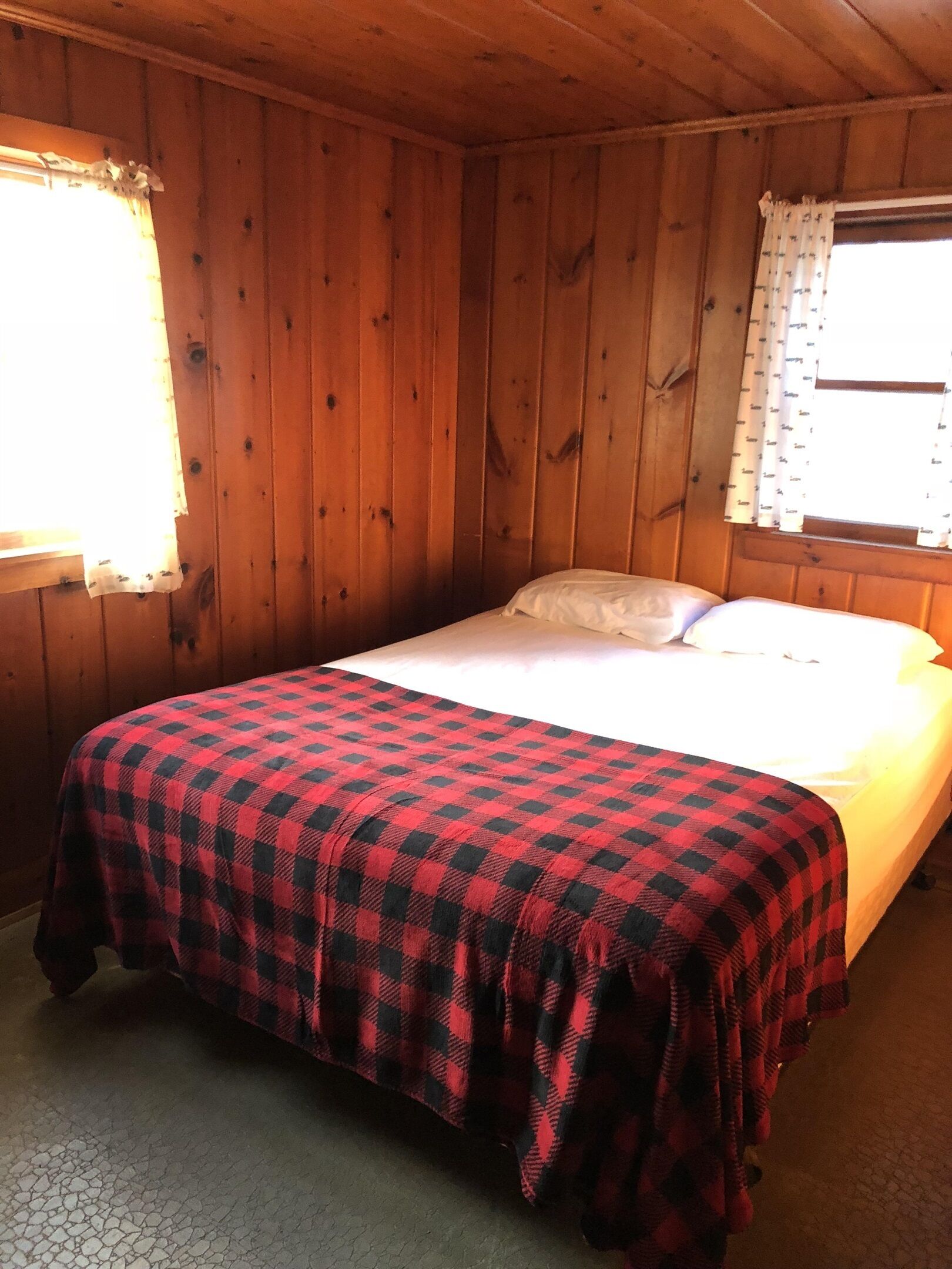 A wooden cabin room with a bed, a window with curtains, and a checkered blanket.