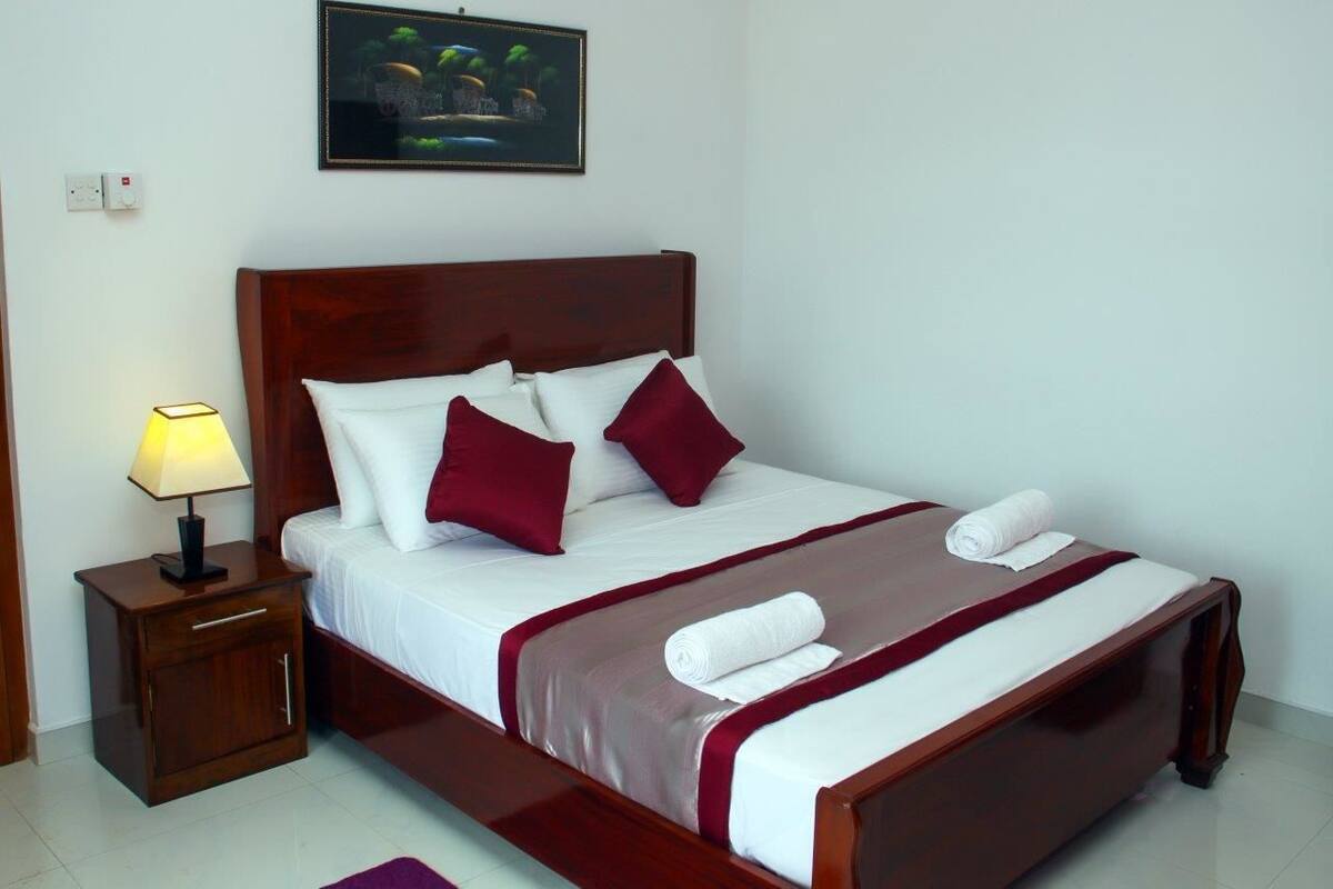 A neatly made bed with white and maroon bedding, a wooden headboard, and a nightstand with a lamp.