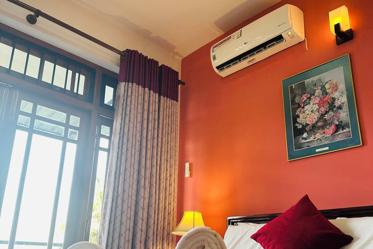 A hotel room with a bed, a window with curtains, an air conditioner, a lamp, and a painting on the wall.