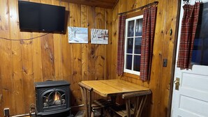 Ein gemütliches Blockhaus-Interieur mit Kaminofen, Holztisch, Wandfernseher und karierten Vorhängen am Fenster.