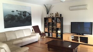 A living room with a beige sofa, a coffee table, a bookshelf, a flat-screen TV, and a large painting of rocks and water. - Free WIFI **200m to beach** R/C Air in all rooms  (Moonta Bay)