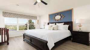 A bedroom with a large bed, two bedside tables with lamps, a ceiling fan, a window with a view, and a decorative mirror on the wall.
