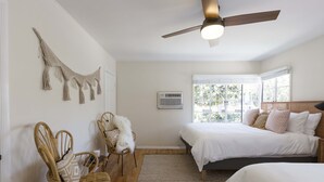 Een slaapkamer met twee bedden, een plafondventilator, een groot raam en een macramé wanddecoratie.