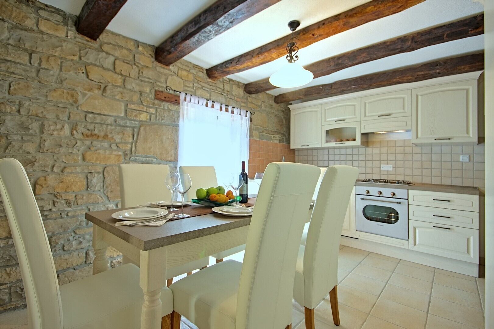 A dining area with a stone wall, wooden beams, white cabinets, and a tiled floor.