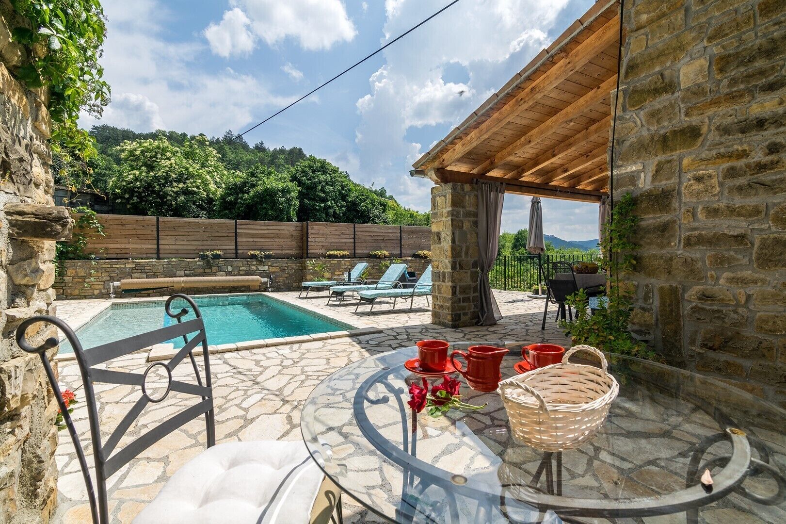 A stone patio with a pool, lounge chairs, a table with red cups and a basket, and a wooden pergola.