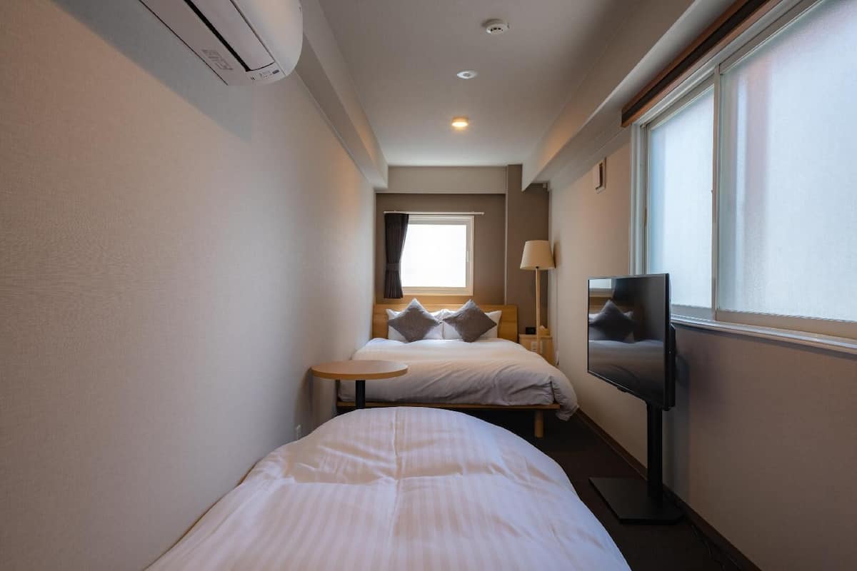 A narrow hotel room with a bed, a small table, a TV, and a window.