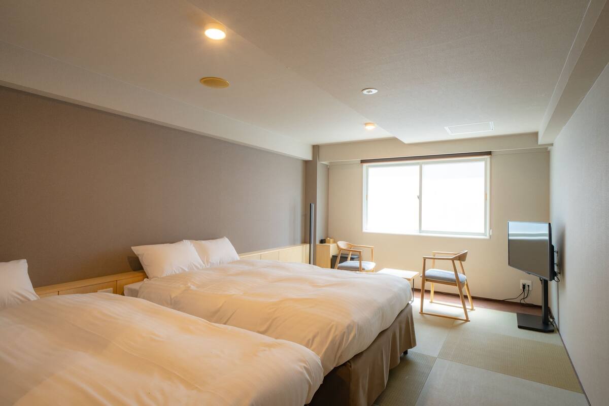 A hotel room with two beds, a desk, a chair, a television, and a large window.