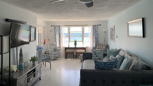 Ein Wohnzimmer mit Deckenventilator, Fernseher, Esstisch mit Stühlen und Meerblick durch große Fenster.