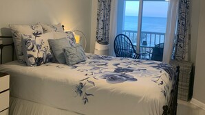 Ein Schlafzimmer mit einem Bett, einem Deckenventilator, einem Fenster mit Vorhängen, einem Sessel und Meerblick.