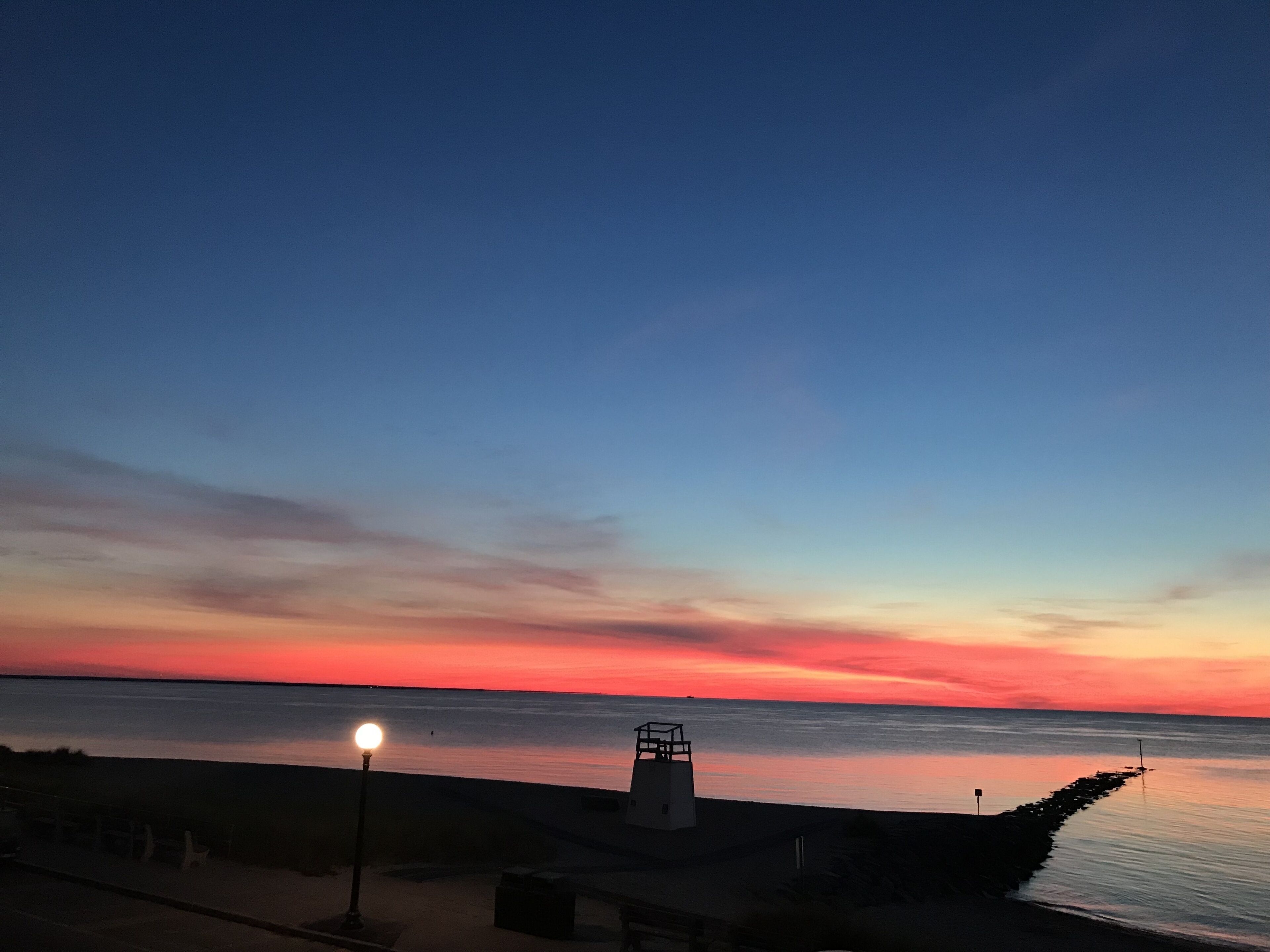 Une vue sur le coucher de soleil en bord de mer, avec un phare, une mer calme et une promenade pavée.