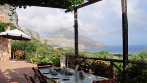 Un espace de restauration en plein air avec une table dressée pour quatre personnes, entourée de chaises en bois, sous une pergola offrant une vue sur la mer et les montagnes.