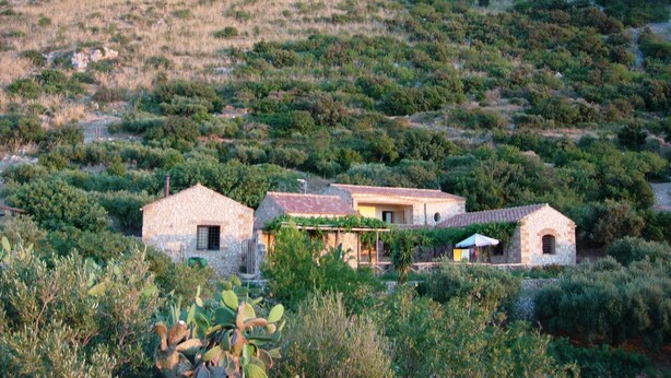 Une maison en pierre avec un toit plat, entourée d’une végétation dense et située au pied d’une colline rocheuse.