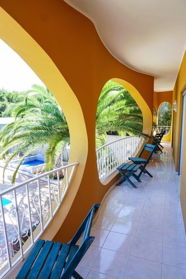 A corridor with arched windows, blue benches, and a pool area visible through the windows.