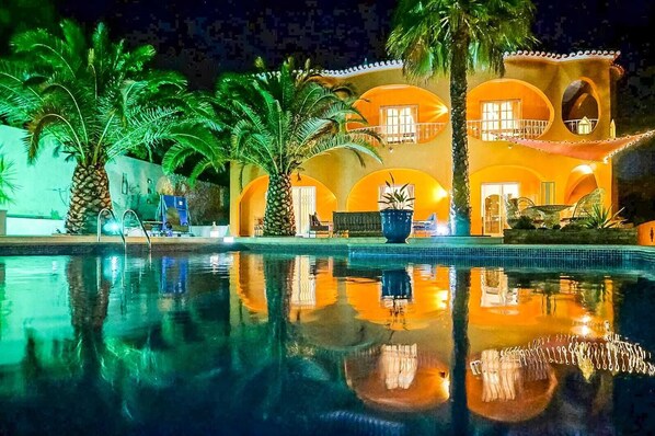 A pool area with a reflection of a building with arched windows and balconies, surrounded by palm trees.