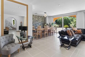 A spacious living room with a grey sofa, a glass coffee table, and a dining area with wooden chairs and a stone wall. - Two Private Pools & Waterfall in a Stunning Portuguese Villa (Armação de Pêra)