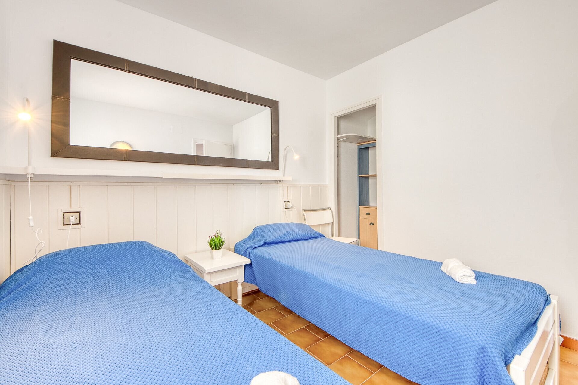 A room with two beds, a small bedside table with a plant, a mirror, and a door leading to another room.