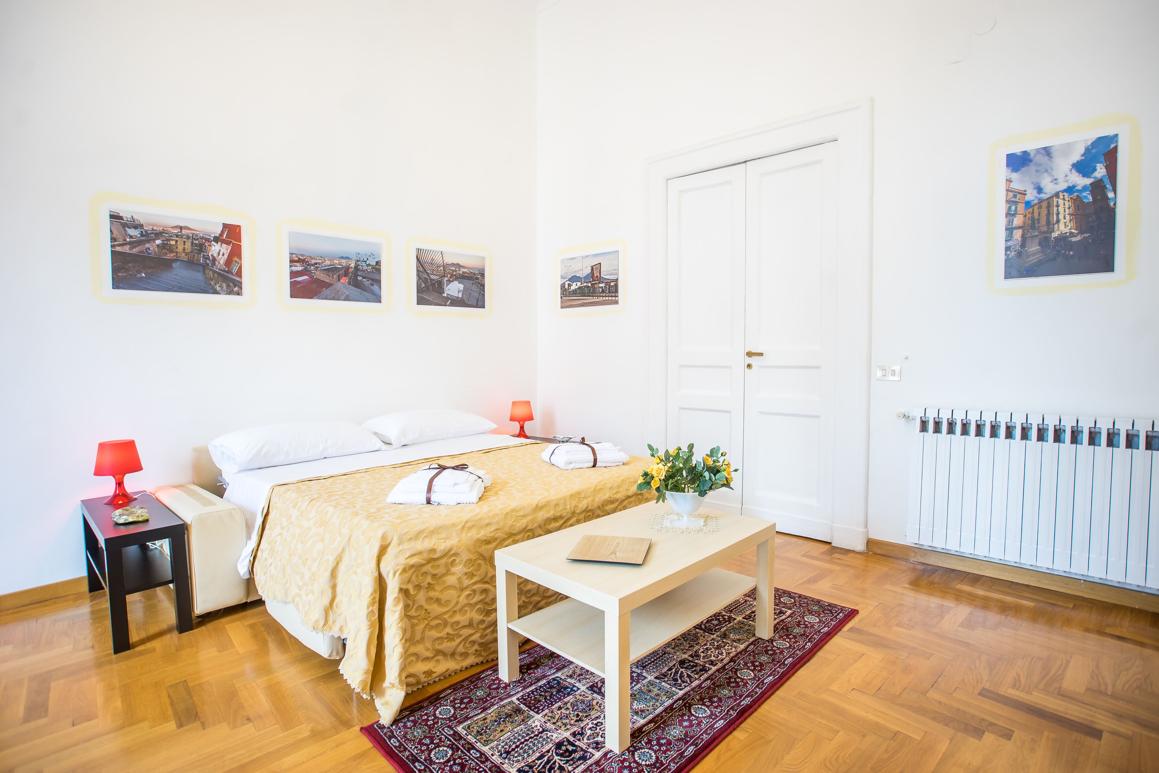 A bedroom with a bed, bedside tables, a desk, a radiator, and framed pictures on the wall.