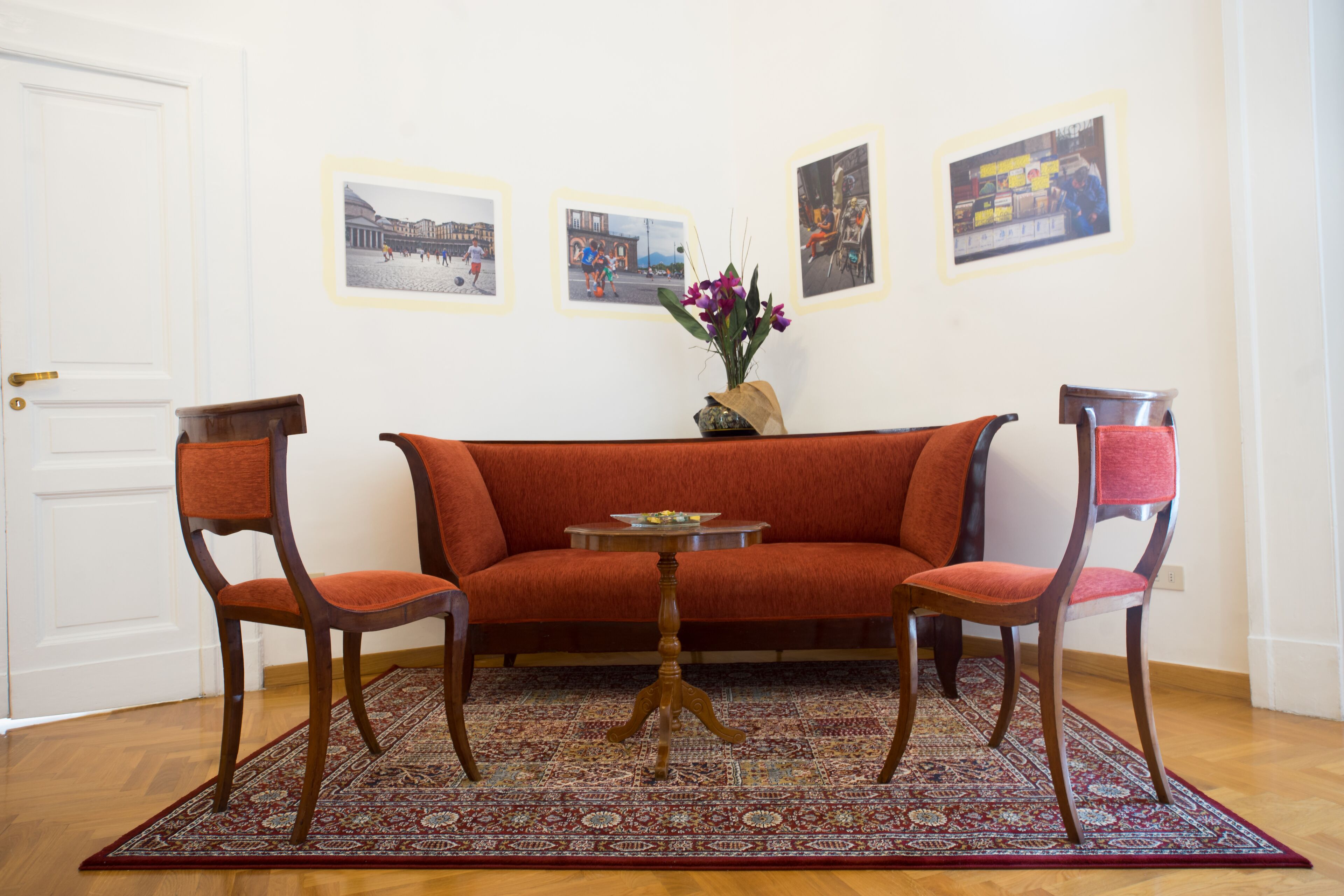 A living room with a red sofa, two wooden chairs, a small table, and a patterned rug. There are three framed pictures on the wall.