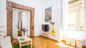 A living room with a wooden floor, a green rug, a white sofa, a small table with a vase of flowers, a flat-screen TV on a shelf, and a window with a view of buildings. - Neapolitan house, unique history and view (Napoli)