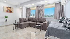 A modern living room with a sectional sofa, a small side table, a decorative plant, and a view of the city through large windows. - Urban Loft with balcony across the Acropolis! (Athina)