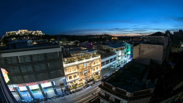 A multi-story building with illuminated signage, a street in front, and a distant hill with lights. - Urban Loft with balcony across the Acropolis! (Athina)