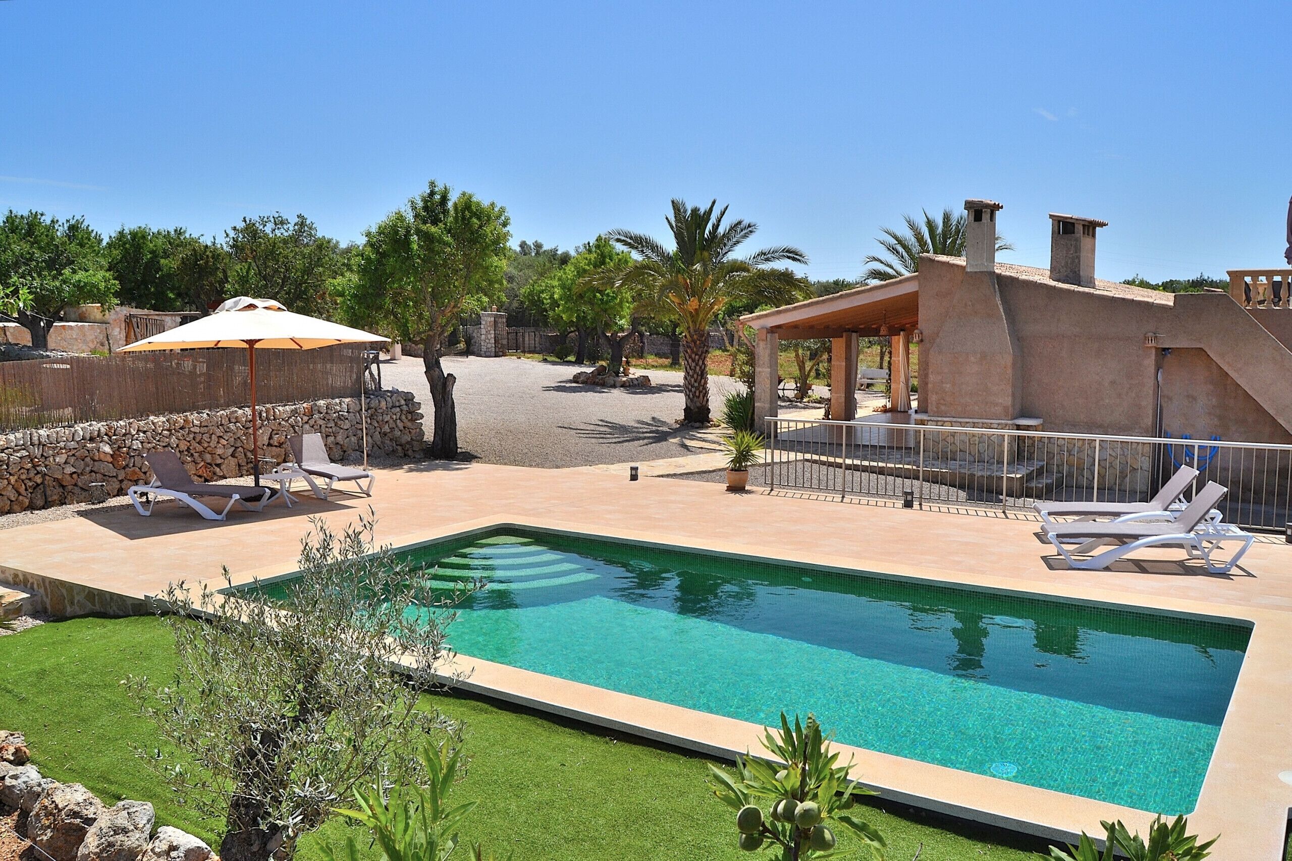 A rectangular pool with clear blue water, surrounded by a stone wall and a grassy area. There are lounge chairs and umbrellas near the pool.