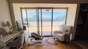 A spacious room with a wooden floor, a desk with a lamp, a rocking chair, a white armchair, and a view through large glass doors.